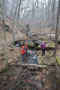 Image of three people collecting water samples from a stream in a wintery forested landscape.