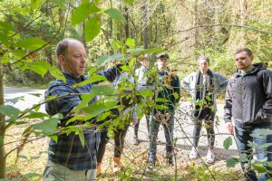 Six people standing in a forest looking at leaves on a budding tree. 