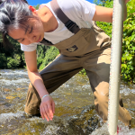 Woman standing in a stream in overalls measuring water flow.
