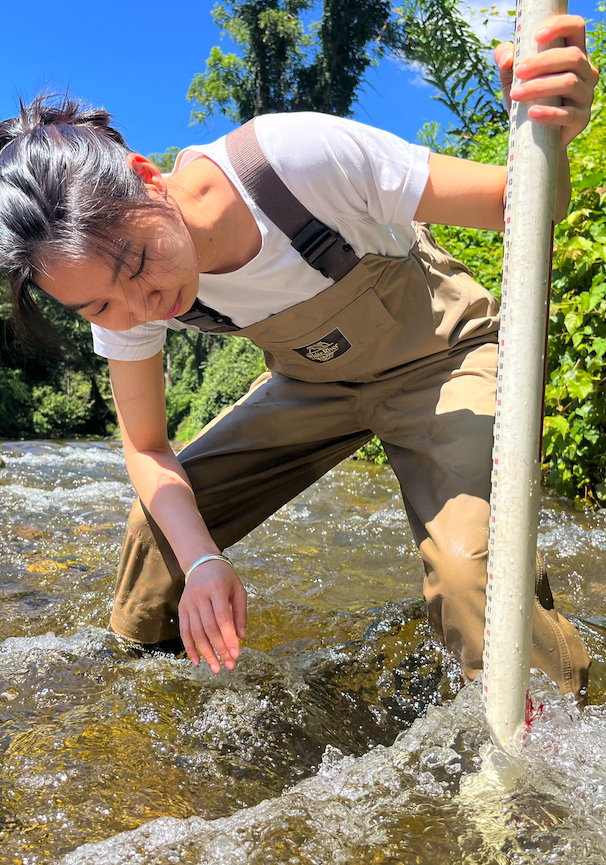 Woman standing in a stream in overalls measuring water flow.
